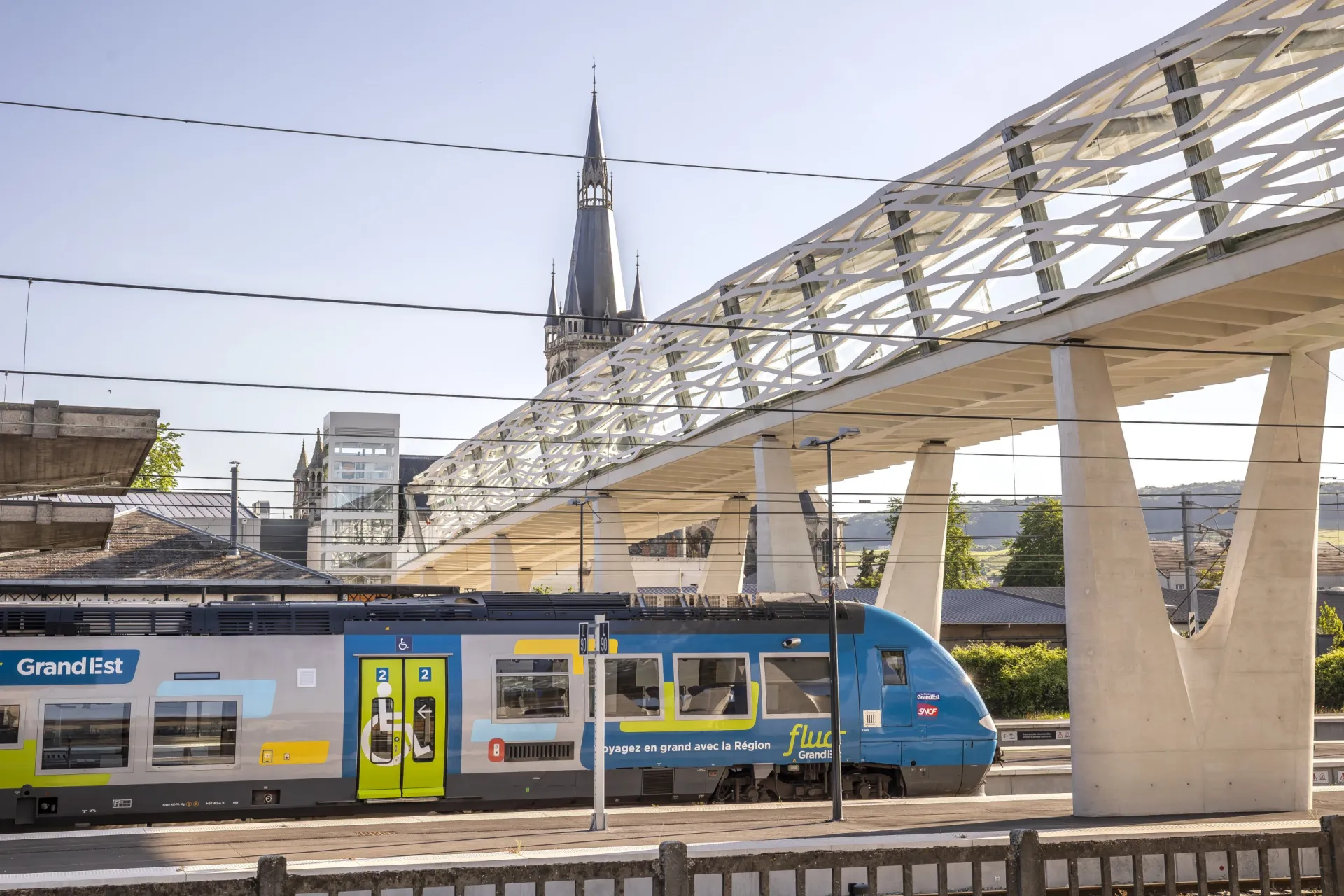Pasarela interbarrios de Épernay de UHPFRC sobre las vías, un tren regional debajo y un campanario al fondo