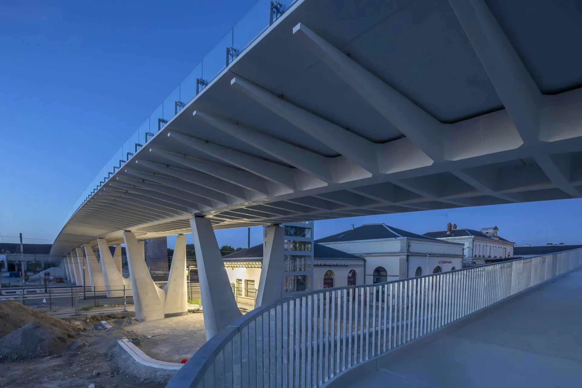 Passerelle interquartier d’Épernay en BFUP à la nuit tombée : sous-face nervurée sur poteaux en V, bâtiments de la gare en fond